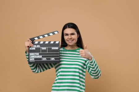 Foto de Feliz actriz con clapperboard mostrando los pulgares hacia arriba sobre fondo beige. Industria cinematográfica - Imagen libre de derechos