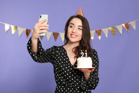 Foto de Fiesta de la mayoría de edad - 21 cumpleaños. Mujer sonriente sosteniendo delicioso pastel con velas en forma de número y tomando selfie contra fondo violeta - Imagen libre de derechos