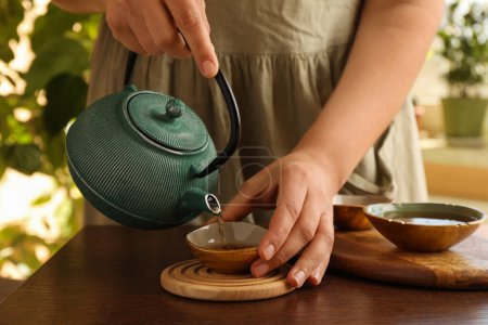Mujer vertiendo té recién hecho de tetera en taza en la mesa de madera en el interior, primer plano. Ceremonia tradicional