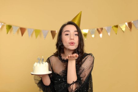 Foto de Fiesta de la mayoría de edad - 21 cumpleaños. Mujer sosteniendo delicioso pastel con velas en forma de número y el envío de beso de aire sobre fondo beige - Imagen libre de derechos