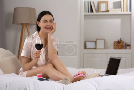 Foto de Hermosa joven con copa de vino dándose pedicura en la cama en casa - Imagen libre de derechos