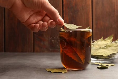 Téléchargez les photos : Femme mettant la feuille de laurier dans une tasse de thé fraîchement infusé à table grise, gros plan - en image libre de droit