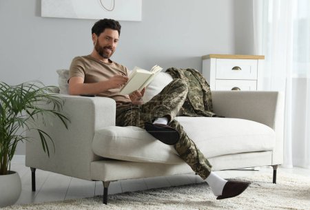 Happy soldier reading book on soft sofa in living room. Military service