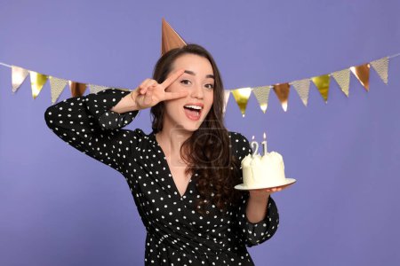 Foto de Fiesta de la mayoría de edad - 21 cumpleaños. Mujer sonriente mostrando signo de paz y sosteniendo delicioso pastel con velas en forma de número sobre fondo violeta - Imagen libre de derechos