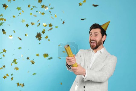 Photo for Emotional man blowing up party popper on light blue background - Royalty Free Image