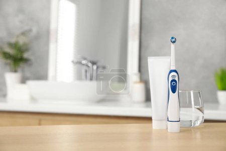 Téléchargez les photos : Brosse à dents électrique, tube avec pâte et verre d'eau sur table en bois dans la salle de bain. Espace pour le texte - en image libre de droit