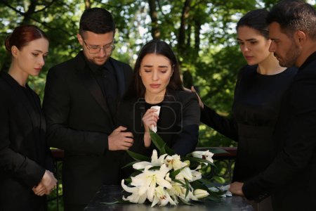 Trauerzeremonie. Trauernde in der Nähe von Granitgrabstein mit weißen Lilien auf Friedhof im Freien
