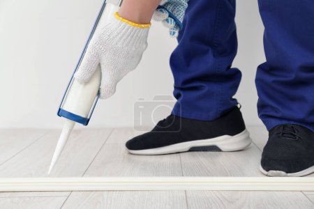 Photo for Man using caulking gun while installing plinth on laminated floor in room, closeup - Royalty Free Image