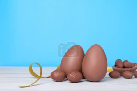 Photo for Delicious chocolate eggs and golden ribbon on white wooden table against light blue background - Royalty Free Image