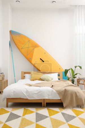 Photo for Large comfortable bed, SUP board and green houseplant in stylish bedroom. Interior design - Royalty Free Image