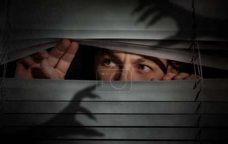 Téléchargez les photos : Worried man looking through window blinds into darkness. Shadow of hands with long claws. Paranoia concept - en image libre de droit