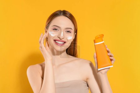 Photo for Beautiful young woman with sun protection cream on her face holding sunscreen against orange background - Royalty Free Image
