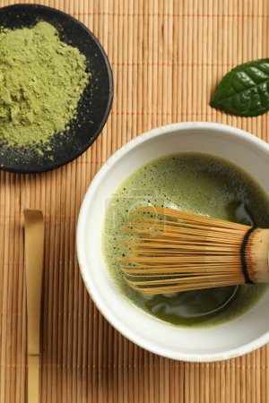 Cup of fresh matcha tea with whisk, spoon and green powder on bamboo mat, flat lay