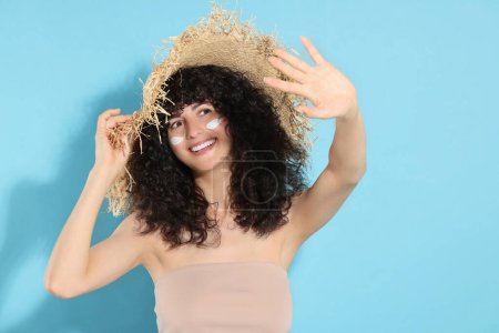 Téléchargez les photos : Belle jeune femme en chapeau de paille avec crème de protection solaire sur le visage sur fond bleu clair - en image libre de droit