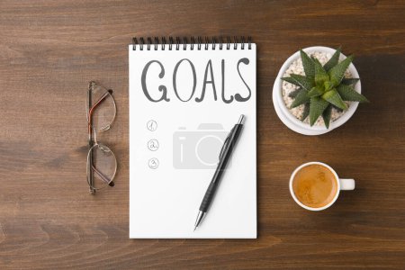 Notebook with empty list of goals, houseplant, glasses, pen and cup of coffee on wooden table, flat lay