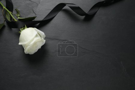 White rose and ribbon on black table, flat lay with space for text. Funeral symbols
