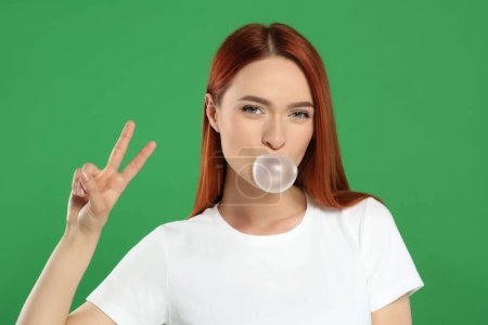 Photo for Beautiful woman blowing bubble gum and showing peace gesture on green background - Royalty Free Image