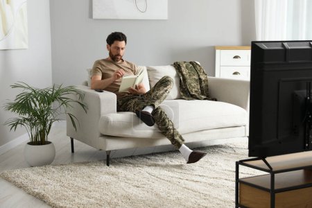 Foto de Soldado leyendo libro sobre sofá blando en sala de estar. Servicio militar - Imagen libre de derechos