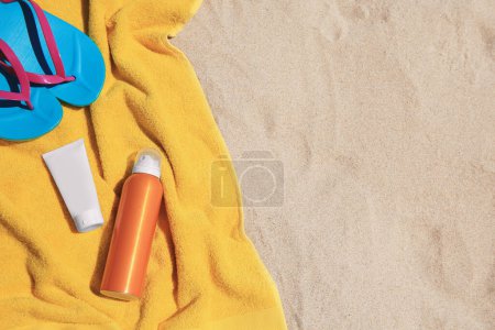 Sunscreens, flip flops and towel on sand, top view with space for text. Sun protection care