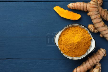 Bowl with turmeric powder and raw roots on blue wooden table, flat lay. Space for text