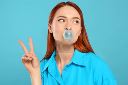 Téléchargez les photos : Belle femme soufflant de la gomme à bulles et montrant un geste de paix sur fond turquoise - en image libre de droit