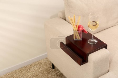 Photo for Glass of white wine and breadsticks on sofa with wooden armrest table in room, space for text. Interior element - Royalty Free Image