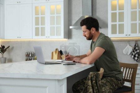 Foto de Soldado tomando notas mientras trabaja con el ordenador portátil en la mesa de mármol blanco en la cocina. Servicio militar - Imagen libre de derechos