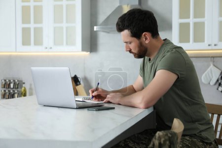Soldier taking notes while working with laptop at white marble table in kitchen. Military service