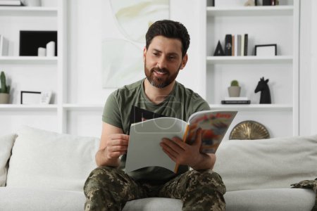 Foto de Feliz soldado leyendo la revista en el sofá de la sala de estar. Servicio militar - Imagen libre de derechos