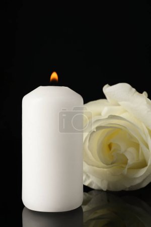 White rose and burning candle on black mirror surface in darkness, closeup. Funeral symbols