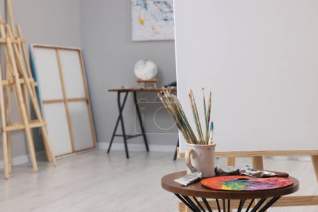 Foto de Caballete de madera con lienzo cerca de pinturas, paleta y pinceles en el estudio del artista, espacio para el texto - Imagen libre de derechos
