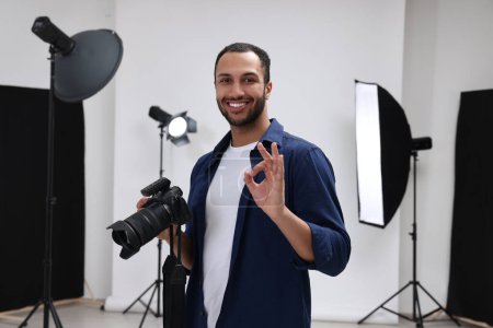 Téléchargez les photos : Jeune photographe professionnel avec appareil photo dans un studio photo moderne montrant OK geste - en image libre de droit