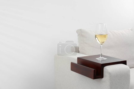 Photo for Glass of white wine on sofa with wooden armrest table indoors, space for text. Interior element - Royalty Free Image