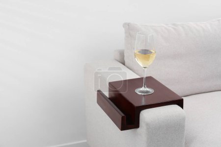 Photo for Glass of white wine on sofa with wooden armrest table indoors, space for text. Interior element - Royalty Free Image