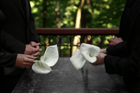 Foto de Personas con flores de lirio de cala cerca de lápida de granito en el cementerio al aire libre, primer plano. Ceremonia funeraria - Imagen libre de derechos