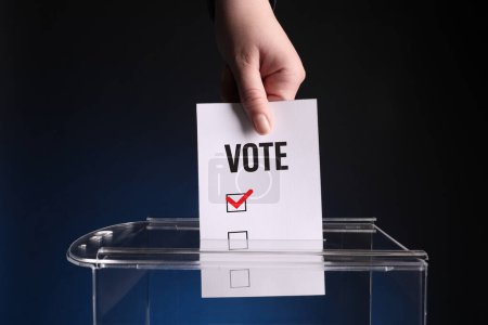 Téléchargez les photos : Femme mettant du papier avec mot Votez et cochez dans l'urne sur fond bleu foncé - en image libre de droit