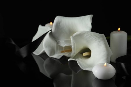 Photo for Burning candles, ribbon and white calla lily flowers on black mirror surface in darkness, closeup. Funeral symbols - Royalty Free Image