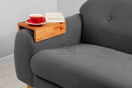 Photo for Cup of tea and book on sofa with wooden armrest table in room. Interior element - Royalty Free Image