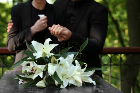 Paar in der Nähe von Granitgrabstein mit weißen Lilien auf dem Friedhof im Freien, selektiver Fokus. Trauerzeremonie