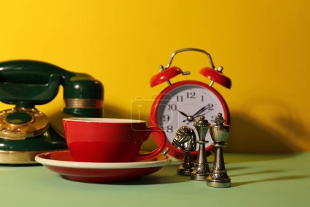Téléchargez les photos : Coupe rouge avec soucoupe, réveil, téléphone vintage et échiquiers sur table vert clair sur fond jaune - en image libre de droit
