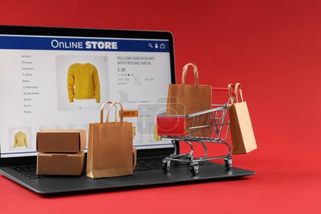 Foto de Tienda online. Ordenador portátil, mini carrito de compras y pequeñas compras sobre fondo rojo - Imagen libre de derechos