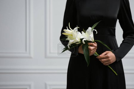 Photo for Woman with lilies near white wall, closeup and space for text. Funeral ceremony - Royalty Free Image