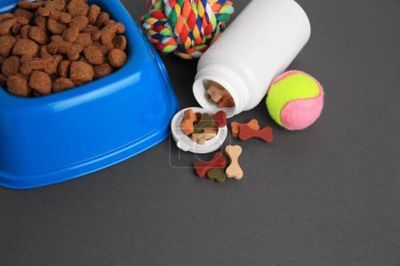 Téléchargez les photos : Bouteille de vitamines, jouets et aliments secs pour animaux de compagnie dans un bol sur fond gris. Espace pour le texte - en image libre de droit