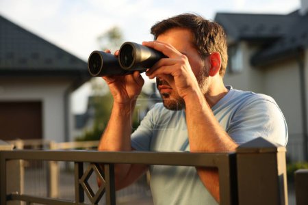 Téléchargez les photos : Concept de vie privée. Homme curieux avec des jumelles espionnant les voisins au-dessus de la clôture à l'extérieur - en image libre de droit