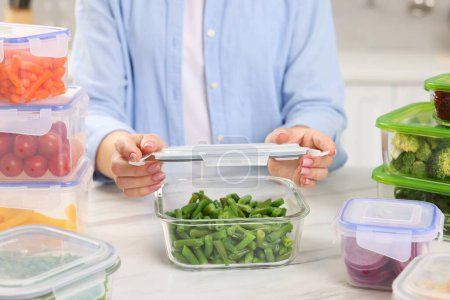 Frau versiegelt Behälter mit grünen Bohnen an weißem Marmortisch in Küche, Nahaufnahme. Lebensmittelaufbewahrung