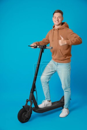 Photo for Happy man with modern electric kick scooter showing thumbs up on light blue background - Royalty Free Image