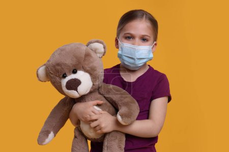 Téléchargez les photos : Jolie fille avec masque médical et ours en peluche sur fond orange - en image libre de droit