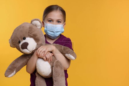 Téléchargez les photos : Jolie fille avec masque médical et ours en peluche sur fond orange, espace pour le texte - en image libre de droit