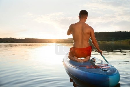 Photo for Man paddle boarding on SUP board in river at sunset, back view - Royalty Free Image