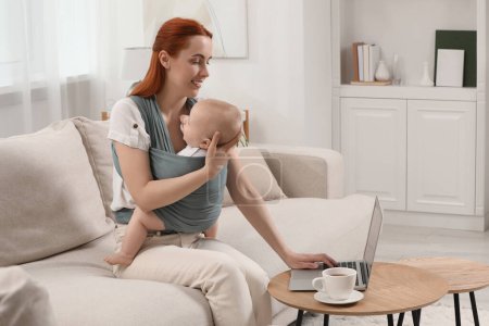 Mutter hält ihr Kind im Tragetuch (Babytrage), während sie zu Hause Laptop benutzt